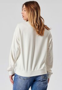 Lichtcreme sweatshirt met een ontspannen pasvorm, ribgebreide manchetten en zoom. Zachte stof; gecombineerd met blauwe denim jeans met een detail op de achterzak.