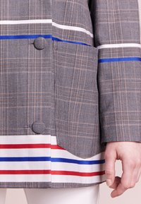 Blazer grigio a quadri con strisce bianche, rosse e blu all'orlo. Presenta una tasca rotonda e un bottone in tessuto come accent. 