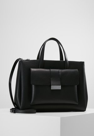 Sort læderhåndtaske med dobbelte håndtag, forflap med metalspænde og aftagelig skulderrem på hvid overflade.