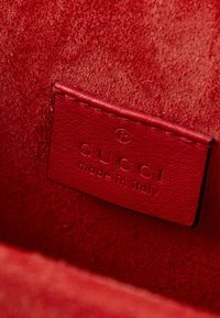 Étiquette en cuir rouge cousue sur du daim rouge avec texte embossé "GUCCI made in Italy" et symbole de marque déposée.
