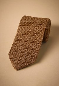 Corbata tejida marrón con un patrón texturizado, que presenta un extremo puntiagudo y un diseño enrollado, sobre un suave fondo beige.