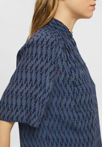Camicia blu navy a maniche corte, con un motivo diagonale di sottili linee arancioni e blu. Include un colletto e una tasca sul petto.