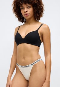 Svart, bøyd bralette med tynne stropper, kombinert med en kremfarget thong med en svart merket linning. Glatt stoff med minimalistisk design.