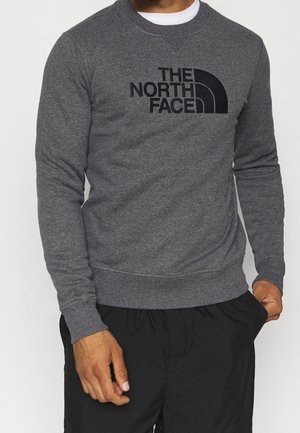 Mężczyzna w ciemnoszarej bluzie z okrągłym dekoltem z czarnym logo "THE NORTH FACE" na piersi, w połączeniu z czarnymi spodniami, stojący na białym tle.