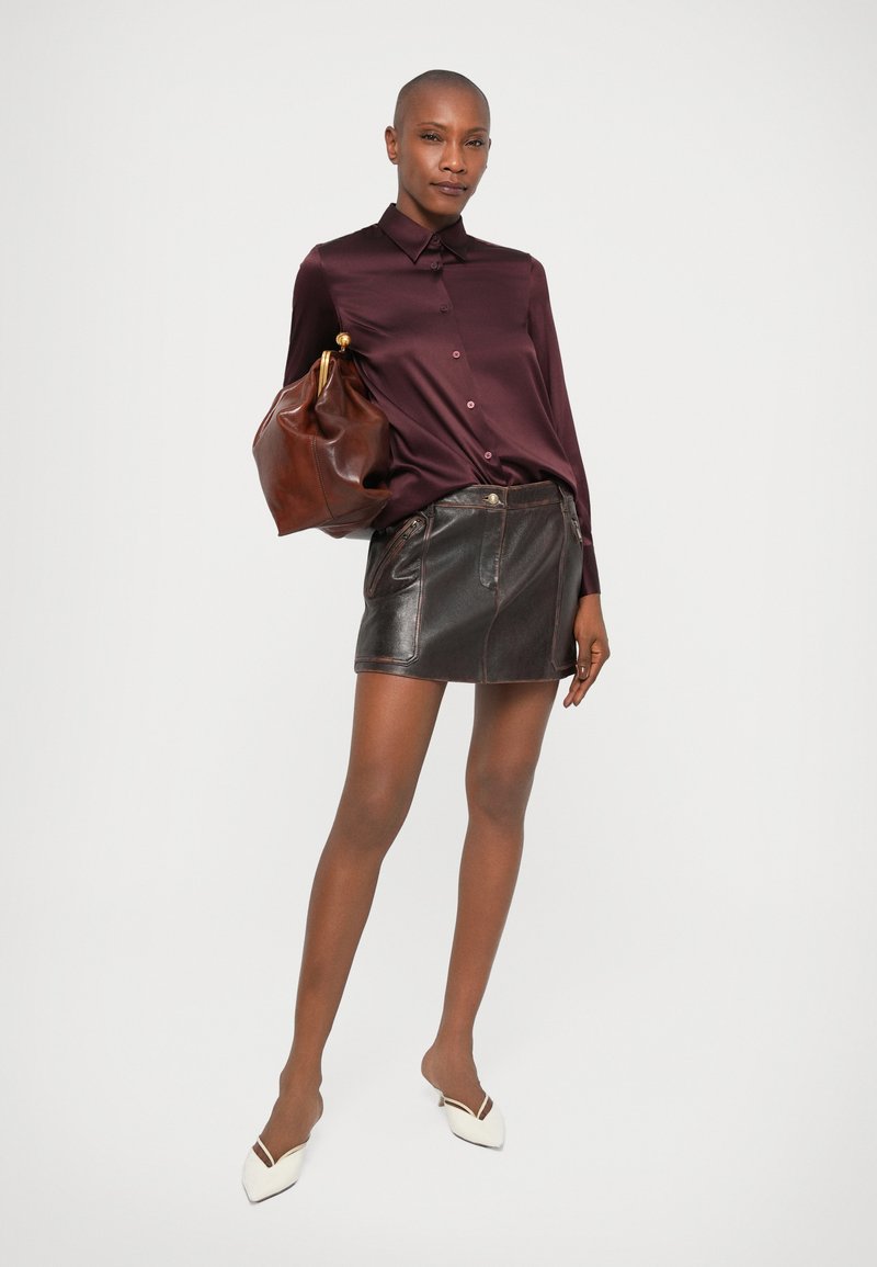 Frau in einem burgunderfarbigen Satin-Hemd mit Knöpfen, dunklem Leder-Minirock, weißen Absätzen, die eine große braune Ledertasche an einem Arm hält.