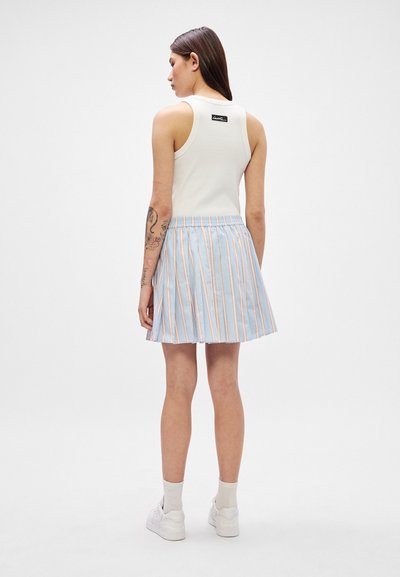Débardeur blanc avec une étiquette de logo noire. Jupe plissée rayée bleu et blanc. Baskets blanches basiques et chaussettes de cheville claires.