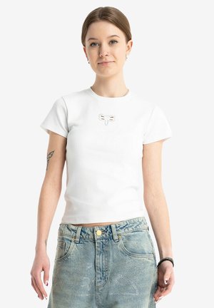 Młoda kobieta w białej bluzce z krótkim rękawem z haftowanym ważką oraz jasnoniebieskich dżinsach z wzorami z kryształków, stojąca na białym tle.