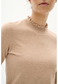 Beige neulepusero, jossa on röyhelöinen korkea kaulus, valmistettu pehmeästä neuloksesta; siinä on kapea malli ja minimimäärä ompelutarkkuuksia.
