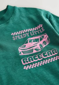 Roheline kapuutsiga särk, millel on roosa võidusõidauto graafika ja tekst "Kiire Väike Võidusõidauto" koos ruudulise musteraktsendiga. Pehme tekstuuri detail on nähtav.