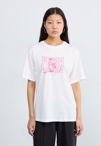 Bílý bavlněný tričko s krátkým rukávem, které má růžový grafický potisk kočky a text "PUMA". Volný střih. Stylizováno s tmavými kalhotami.