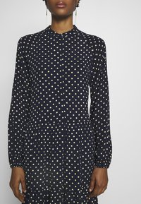 Femme portant une robe à manches longues bleu marine avec des pois jaune clair, debout devant un fond uni gris clair.