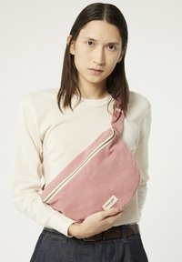 Sac à bandoulière en toile de couleur rose doux avec fermeture éclair dorée et étiquette de marque. Forme incurvée et sangle réglable pour un port polyvalent.