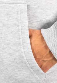 Tejido de sudadera gris con una superficie texturizada. Una mano está colocada en un bolsillo lateral. Los detalles de los puños son acanalados y las costuras son visibles.