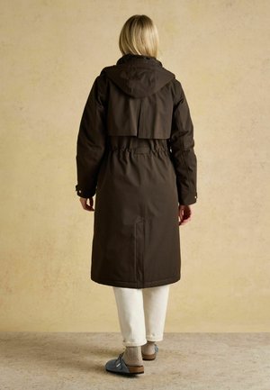 Woman wearing a dark brown hooded long coat, white pants, beige socks, and blue slip-on shoes standing on a beige floor against a beige wall.