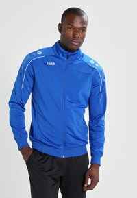 Blauwe sportjas met een volledige ritssluiting, gemaakt van een glad materiaal. Bevat witte accenten en vijf ronde knopen langs de schouders.
