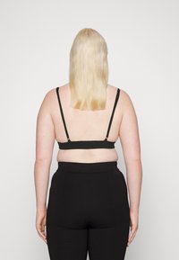Mustat bralette-hihnat, joissa on säädettävät hihat, sileä kangas ja minimalistinen muotoilu; yhdistettynä korkeavyötäröisiin mustiin housuihin, nähtynä takaa.