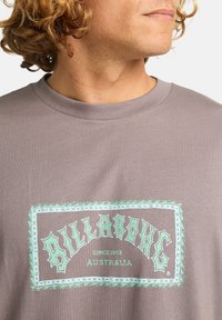 Šedé bavlněné tričko s velkým tyrkysovým grafickým designem a textem "BILLABONG AUSTRALIA" a dekoračním okrajem. Kulatý výstřih.