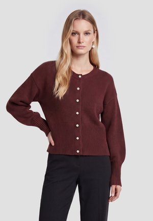 Lyshåret kvinde iført en vinrød cardigan med perleknapper, sorte bukser og perle-øreringe, der står foran en grå baggrund.