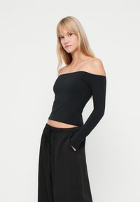 Μαύρη μπλούζα με ανοιχτούς ώμους και μακριά μανίκια, φτιαγμένη από μαλακό ύφασμα, εφαρμοστή σχεδίαση, cropped μήκος, συνδυασμένη με φαρδιά μαύρα παντελόνια με πλευρικές τσέπες.