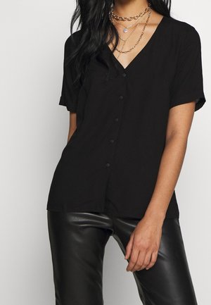 Vrouw draagt een zwart kort mouws blouse met knopen, zwarte leren broek en gelaagde gouden kettingen met een hanger.
