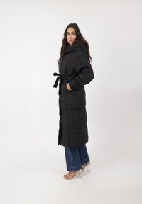Abrigo largo acolchado negro con capucha, cintura ceñida y dos bolsillos laterales. Combinado con jeans acampanados y sandalias con tacón transparente.