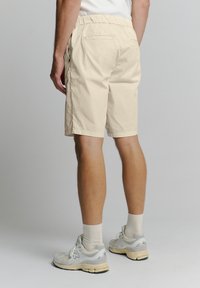 Beige shorts met een elastische tailleband, voorzakken en twee achterzakken. Draag ze met grijze sportschoenen en lichte sokken.