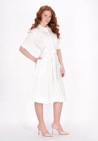 Weißes Hemdkleid mit kurzen Ärmeln, tailliert mit einem Gürtel, verfügt über eine durchgehende Knopfleiste und einen ausgestellten Rock. Kombiniert mit hautfarbene High Heels.