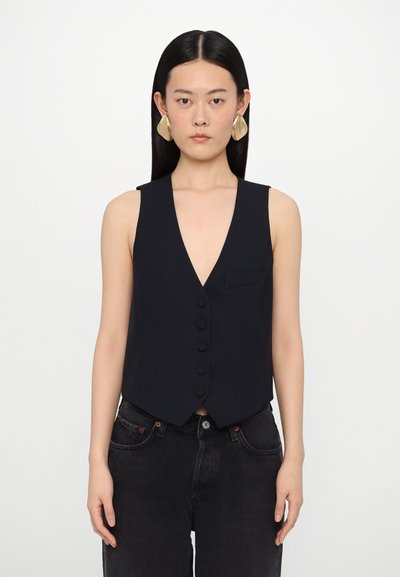 Gilet senza maniche blu navy con scollo a V profondo, chiusura a cinque bottoni e una piccola tasca sul petto, realizzato in tessuto liscio. Indossato con jeans neri.