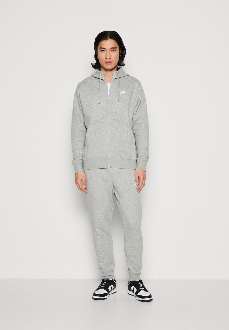 Grau zip-up Kapuzenpullover und passende Jogginghose aus weichem Stoff, mit einer Fronttasche und Logodetails. Schwarze und weiße Sneakers runden den Look ab.