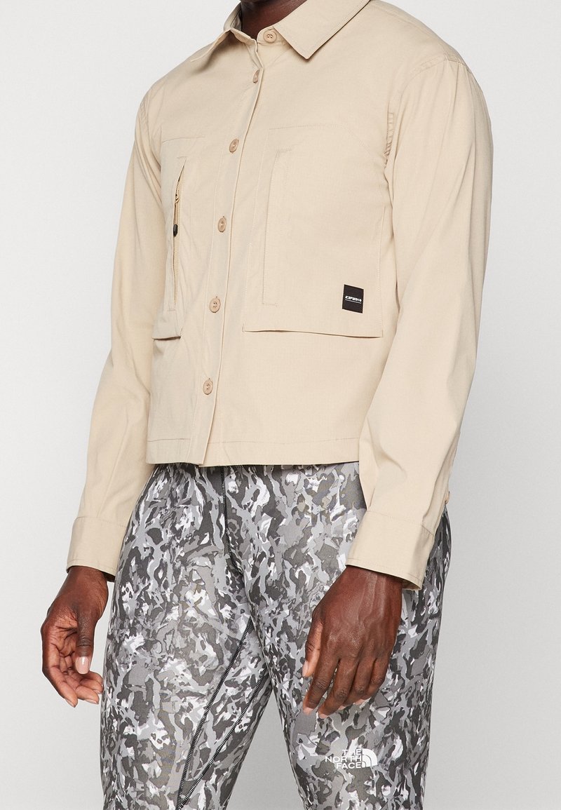 Giacca beige con bottoni, due tasche frontali, tessuto leggero e dettagli di cucitura visibili, abbinata a pantaloni grigi con motivo camouflage.