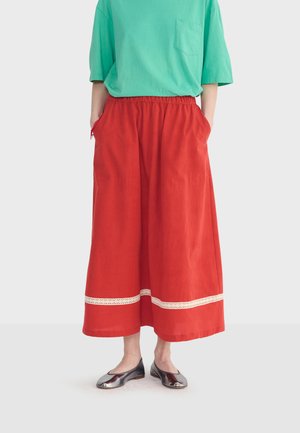 Osoba w zielonkawoniebieskiej koszulce z krótkim rękawem, czerwonych szerokich spodniach z wysokim stanem i białym obszyciem przy nogawce oraz w błyszczących srebrnych baletkach, z rękami w kieszeniach.