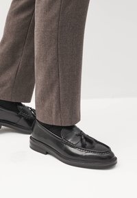 Les mocassins en cuir noir présentent un design tressé et des accents de glands, associés à un pantalon texturé marron clair.