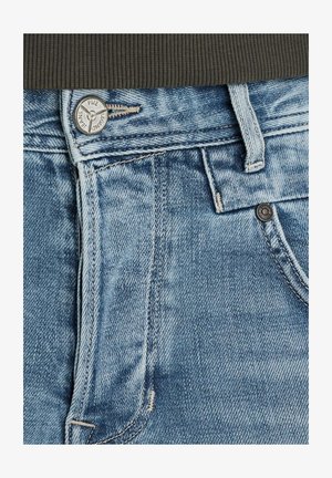 Svijetloplave traperice imaju metalnu kopču, петље за pojas i kontrastne šavove. Prisutna je vidljiva tekstura i lagano izblijeđivanje.