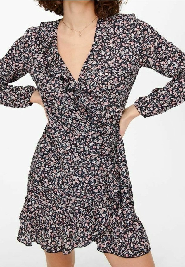 Femme portant une robe portefeuille noire à manches longues avec des fleurs roses et blanches, mains sur les hanches, et un décolleté en V avec un détail volanté.