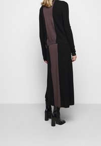 Kvinna som bär en svart och brun färgblockerad långkjol och matchande tröja, i kombination med svarta klackstövlar, stående med ryggen mot kameran.