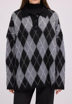 Femme portant un pull ample à motif losanges noir et gris avec un col noir et trois boutons, sur un pantalon noir.