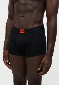 Torse masculin et jambes supérieures portant un boxer noir Hugo avec un patch logo orange sur la ceinture, sur un fond uni.