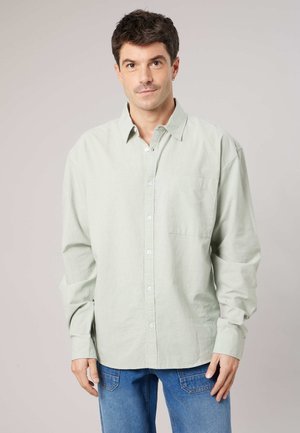 Homme aux cheveux courts et foncés portant une chemise boutonnée à manches longues vert pâle et un jean bleu, debout devant un fond neutre.