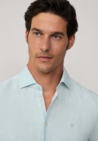 Light blue linen shirt with a button-down collar, subtle texture, and a small embroidered logo on the chest.