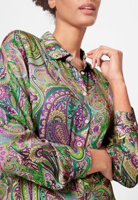 Kleurige paisley-blouse in groen, paars en crèmekleurig. Heeft een kraag, knopen en een zijdezachte textuur met ingewikkelde bloempatronen.