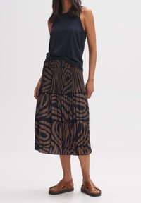 Kvinna iklädd en svart ärmlös topp, brun och svart abstrakt mönstrad midiklänning och bruna plattformssandaler stående mot en vit bakgrund.