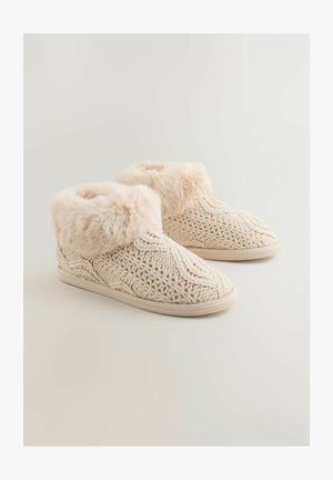 Beige strikkede hjemmesko med en blød kunstig pelskant, tekstureret design og en flad skridsikker sål for komfort og stabilitet.