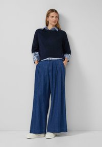 Navy gebreide trui over een blauw-wit gestreept shirt, gecombineerd met losse, geplooide donkerdenim broek en witte sneakers.