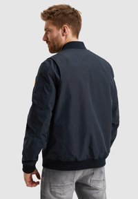 Chaqueta bomber azul marino con cuello y puños acanalados, que presenta una textura suave y una insignia de logo en la manga izquierda.