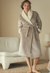 Accappatoio color taupe realizzato in morbido tessuto peluche con un collo e una cintura in pelliccia sintetica color crema, dotato di due tasche laterali e lungo fino a metà polpaccio.