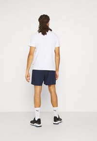White short-sleeve T-shirt paired with navy shorts, black athletic shoes, and white socks with a black logo, viewed from the back.