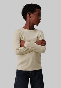 Camicia di cotone beige a maniche lunghe con collo tondo e piccolo logo ricamato sul petto, abbinata a jeans in denim scuro.