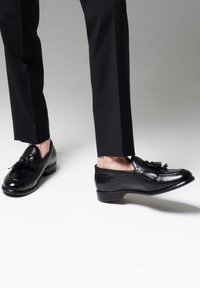 Czarne skórzane mokasyny o gładkim wykończeniu, z frędzlami jako akcentem. Buty mają zaokrąglony czubek i niski obcas, zestawione z czarnymi spodniami.