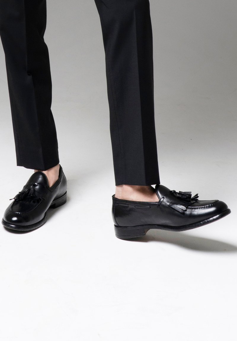 Czarne skórzane mokasyny o gładkim wykończeniu, z frędzlami jako akcentem. Buty mają zaokrąglony czubek i niski obcas, zestawione z czarnymi spodniami.