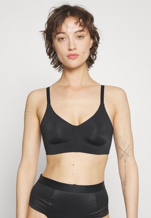 Frau mit kurzen lockigen Haaren, die einen schwarzen BH und hoch taillierte Unterwäsche trägt, vor einem schlichten weißen Hintergrund stehend.